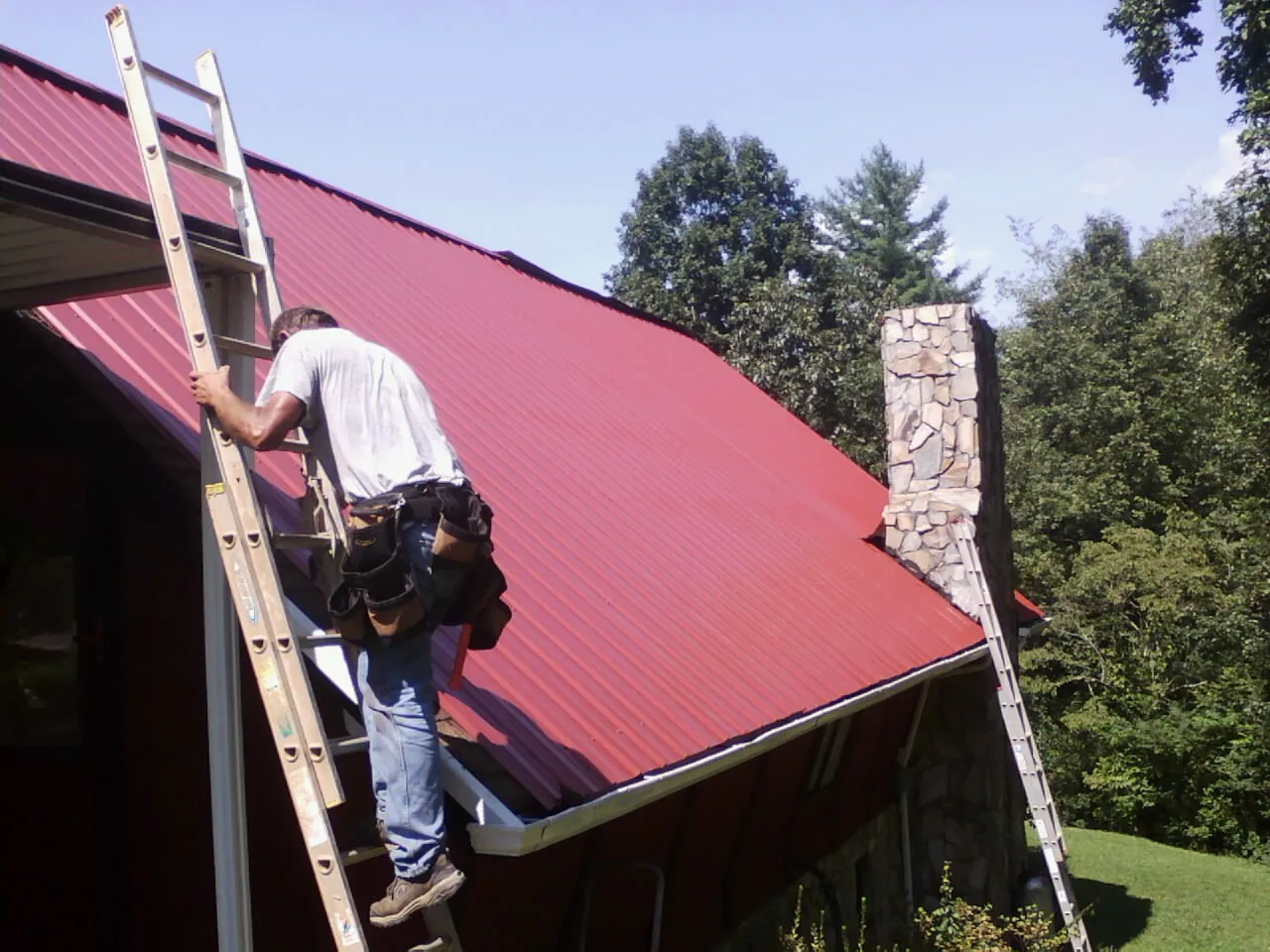 Roofer on a ladder beside a red metal roof and stone chimney — Soffit & Fascia Repair by Roofing Brothers in Saint Benedict